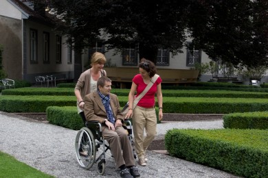 Zwei Frauen schieben älteren Mann im Rollstuhl durch einen Park